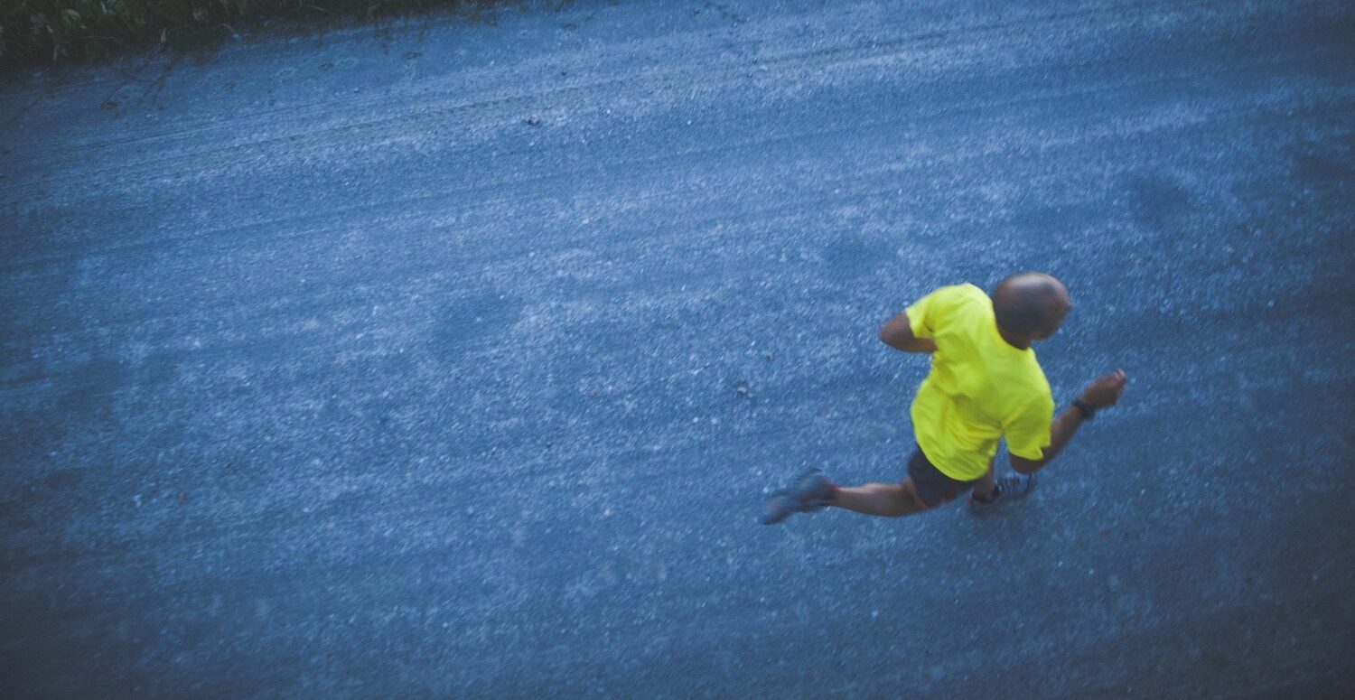 Person in bright yellow athletic shirt running on a blue-tinted gravel path with grass along the edge beyond the road edge.
