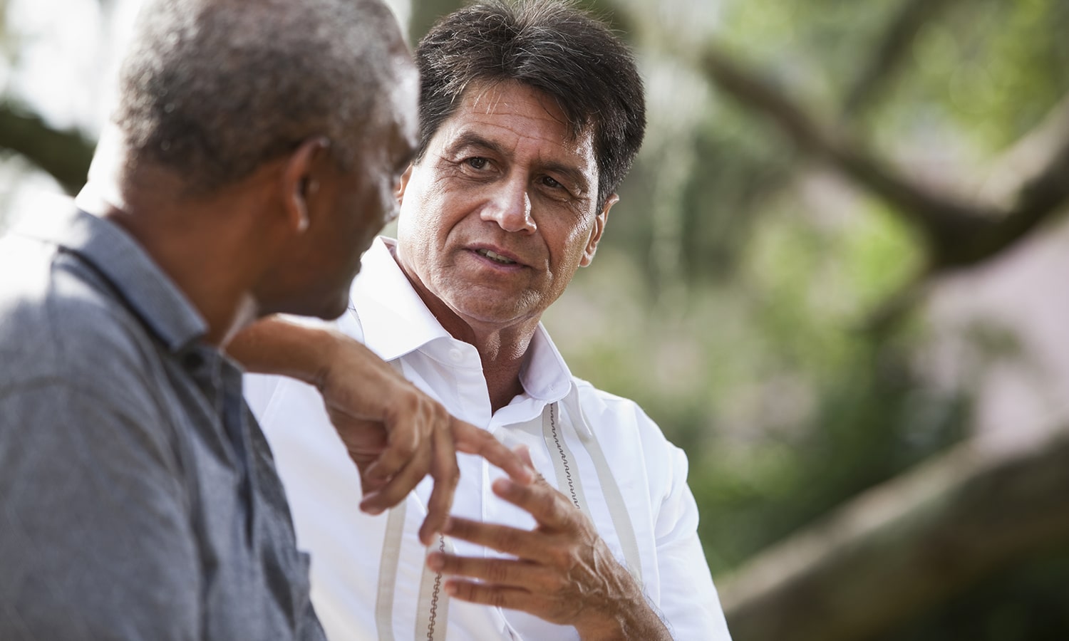 Multi-ethnic men talking Two men in an outdoor setting converse; the man in a white shirt with suspenders gestures as he speaks to another man in a gray shirt.
