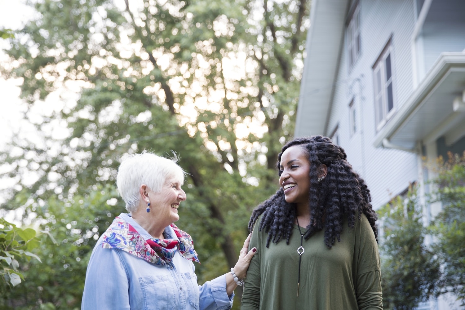 walk_together Two women chatting and smiling in a yard beside a light-blue house, with green trees in the background.