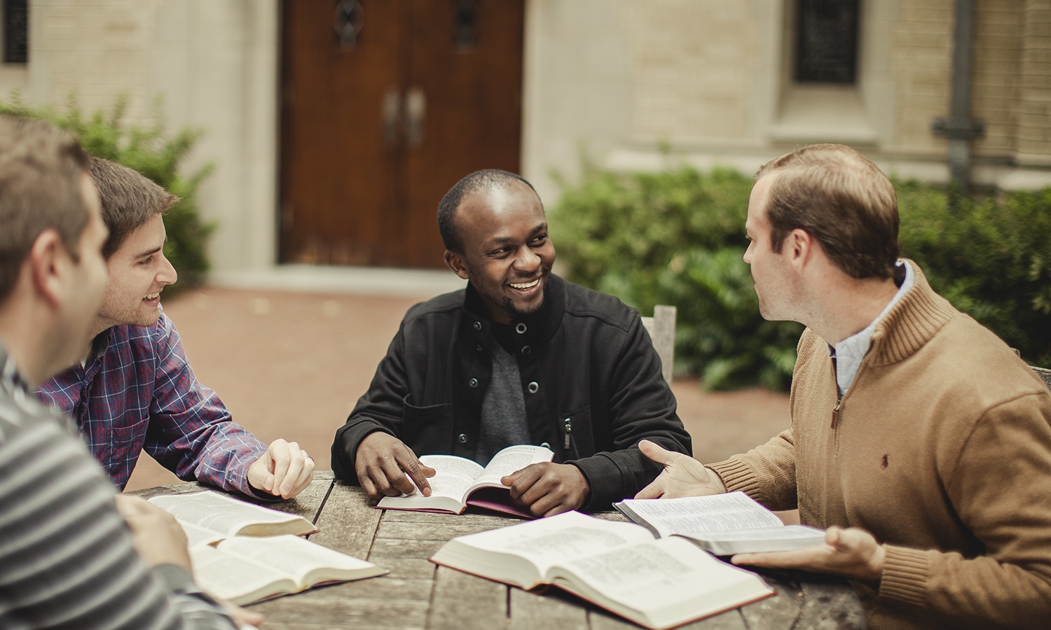 Four people sit around a wooden table outdoors, smiling and studying with open books in front of them.