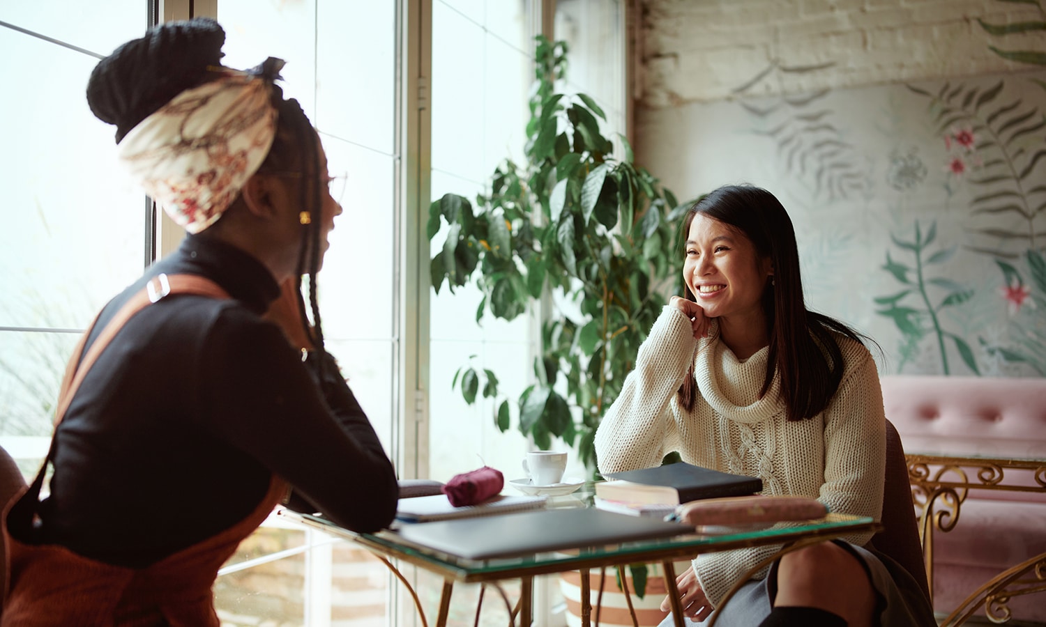Two happy multicultural female college girls sit in the cafeteria chatting and gossiping Female students and girls talk Two women sit at a glass table by a large window, smiling as they talk.