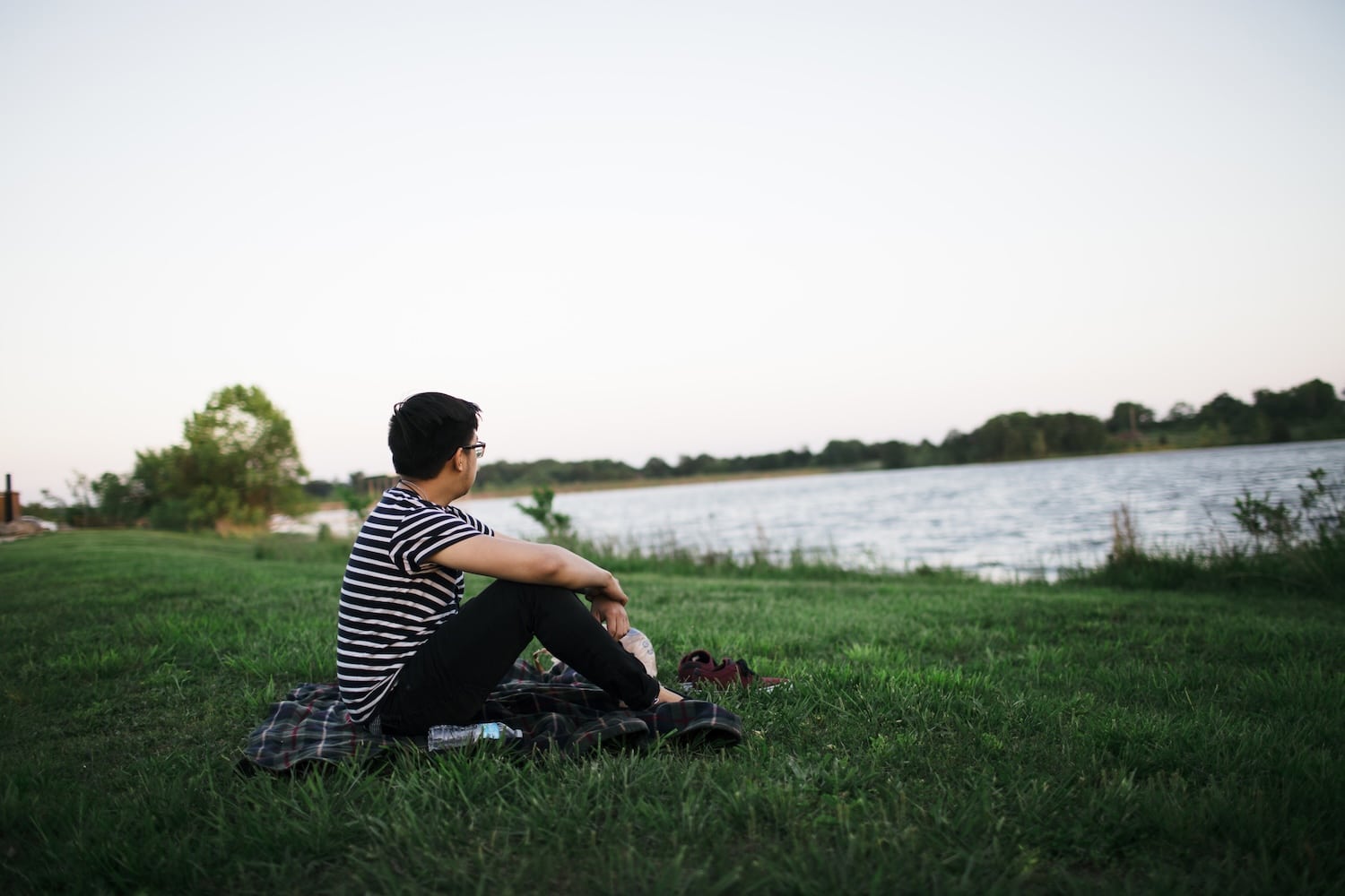Man sitting on a plaid blanket by a lake, gazing at the water