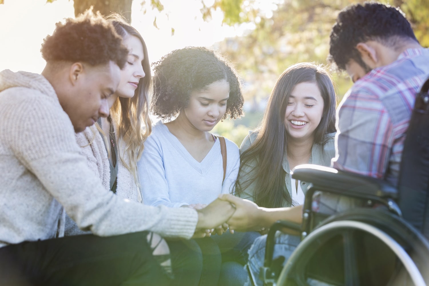 Group of diverse young adults outdoors, joining hands in a circle with a person in a wheelchair present, showing support and teamwork.