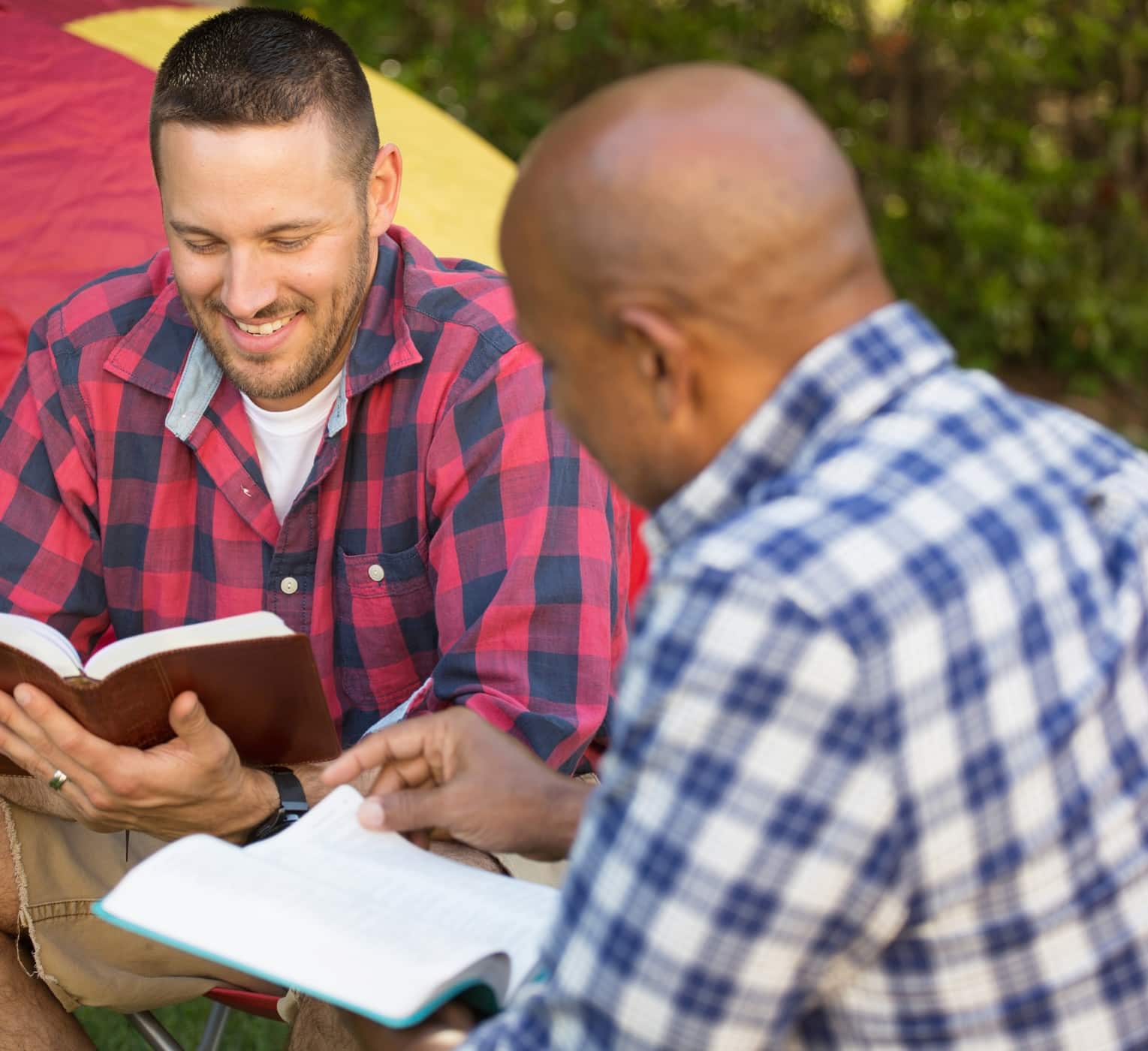 Men having a bible study on a camping trip Two men sitting outdoors and smiling as they read and discuss open Bibles together, illustrating Christian fellowship, Bible study, and discipleship.